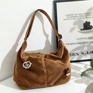 Könnte beinhalten: Braune Cord-Umhängetasche mit runder Form. Die Tasche hat einen langen Riemen und ist mit einem herzförmigen Aufnäher und einem kleinen Teddybären verziert. Die Tasche steht vor einem gerahmten Poster.