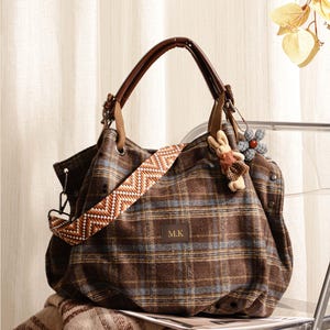 Könnte beinhalten: Eine braune karierte Tragetasche mit einem braunen Ledergriff und einem gemusterten Schultergurt. Die Tasche hat einen kleinen Hasen- und Blumenanhänger. Die Initialen "M.K" sind auf der Vorderseite der Tasche abgebildet.