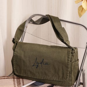 Op de afbeelding: Een olijfgroene canvas schoudertas met een overslagflap waarop de naam Lydia in zwarte cursieve letters is geborduurd. De tas is voorzien van een brede schouderband en zijvakken, gemaakt van duurzame stof met zichtbare stiksels.