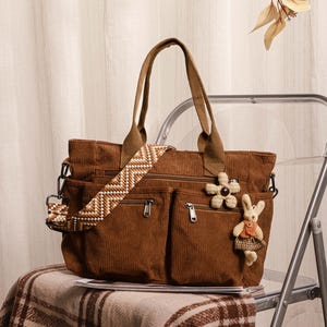 Puede incluir: Un bolso tote de pana marrón con múltiples bolsillos exteriores y una correa para el hombro estampada. La bolsa presenta un charm de flor y conejo. Tiene dos asas superiores y está sobre una manta a cuadros.