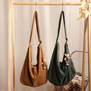 May include: Two slouchy corduroy hobo bags hanging from a wooden rack. One bag is tan and the other is dark green. Each bag features a long shoulder strap and a small, plush rabbit toy attached to the side, wearing a plaid dress and brown vest.