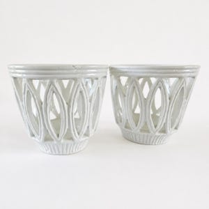 May include: Two white ceramic bowls featuring a decorative openwork lattice design with repeating oval cutouts. Each bowl has a smooth, glossy finish, a flared rim, and a textured base with vertical ridges, creating a uniform appearance.