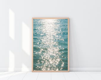 May include: A framed print featuring a photograph of ocean water shimmering with sunlight. The water is a mix of blue and turquoise, with bright, sparkling highlights. The frame is a light wood color, and the print is displayed against a white wall and floor.