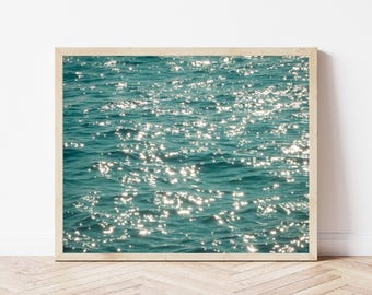 May include: A framed print featuring a close-up of ocean water. The water is a teal color, with small waves and sparkling highlights from the sun. The frame is a light wood color, and the print is displayed against a white wall.