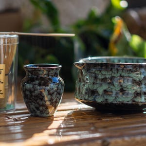 Puede incluir: Un juego de té matcha con un recipiente transparente con un batidor de bambú, un jarrón pequeño y oscuro y un cuenco texturizado esmaltado en turquesa. El jarrón y el cuenco tienen un patrón moteado. El recipiente tiene texto japonés.