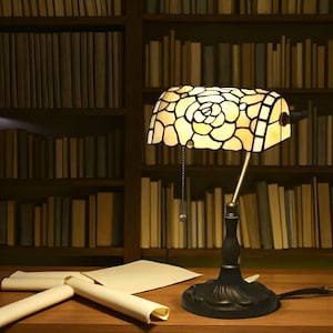 May include: A desk lamp with a stained glass shade featuring a floral pattern. The lamp has a bronze base and an adjustable arm. The background shows a wooden bookshelf filled with books and some rolled-up papers.