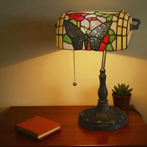 May include: A stained glass desk lamp with a butterfly design. The lamp features a rectangular shade with a colorful floral and butterfly motif. The base is dark metal, and a small succulent plant sits nearby.