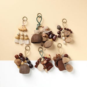 May include: A collection of six decorative keychains with various designs. Each keychain features a gold-colored ring and is adorned with natural elements like nuts, beads, and tassels in shades of brown, beige, and cream. The background is a soft yellow.