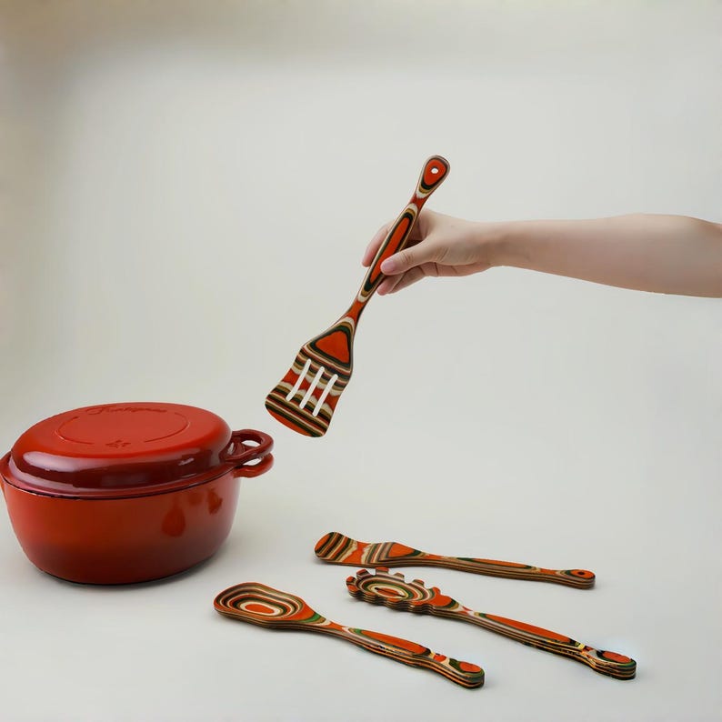 May include: A red enameled cast iron Dutch oven with a lid, and a set of wooden kitchen utensils. The utensils include a slotted spatula, a spoon, and two other cooking tools with orange, brown, and white layered designs.
