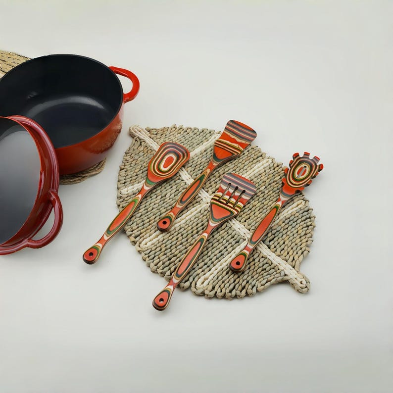 May include: A set of five colorful wooden kitchen utensils, including a spoon, spatula, fork, and two other tools, arranged on a woven placemat. A red cast iron pot and lid are in the background. The utensils have a layered, striped design.