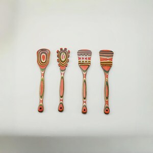 May include: Four wooden kitchen utensils with red, orange, and green accents. The set includes a spoon, a slotted spoon, and two spatulas. Each utensil has a unique design on the head and a long handle.