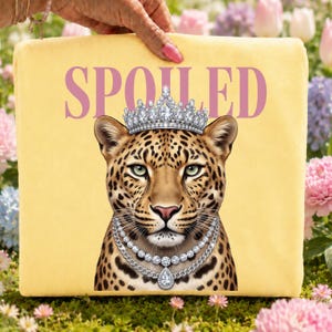 May include: A square yellow pillow featuring a graphic of a leopard wearing a silver crown and a layered diamond necklace. The word SPOILED is printed in large pink capital letters above the leopard's head. The design is centered on the pillow.
