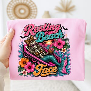 Op de afbeelding: Roze t-shirt met een kleurrijke afbeelding. De afbeelding toont een skeletzeemeermin in een kist, met een zonnebril en een luipaardstrik. De tekst luidt "Resting Beach Face". Tropische bloemen en een zonsondergang zijn ook inbegrepen.