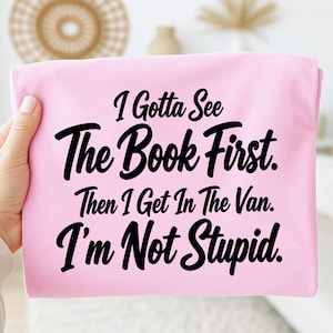 May include: A light pink t-shirt featuring black text that reads: I Gotta See The Book First. Then I Get In The Van. I'm Not Stupid. The shirt is made of a soft fabric and displays the message in a bold, black, cursive and print font combination.