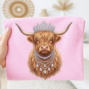 May include: A light pink t-shirt featuring a graphic illustration of a brown Highland cow wearing a large, ornate silver crown encrusted with clear crystals and a multi-layered silver necklace with dangling teardrop-shaped crystal pendants.