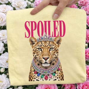 Op de afbeelding: Een lichtgeel t-shirt met een afbeelding van een luipaard die een met juwelen bezette tiara en een meerlaagse edelstenen ketting draagt. Het woord SPOILED staat in grote, dikke roze hoofdletters boven de kop van de luipaard gedrukt.