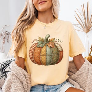Puede incluir: Una camiseta de manga corta de color amarillo pálido con un gran gráfico de una calabaza de patchwork. El diseño de la calabaza está compuesto por varios patrones de tela, incluyendo cuadros vichy, flores y lunares en tonos naranja, verde y crema.