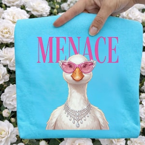 May include: A light blue towel with the word "MENACE" in pink. The towel features a cartoon goose wearing pink sunglasses and a diamond necklace. The goose has an orange beak and white feathers.