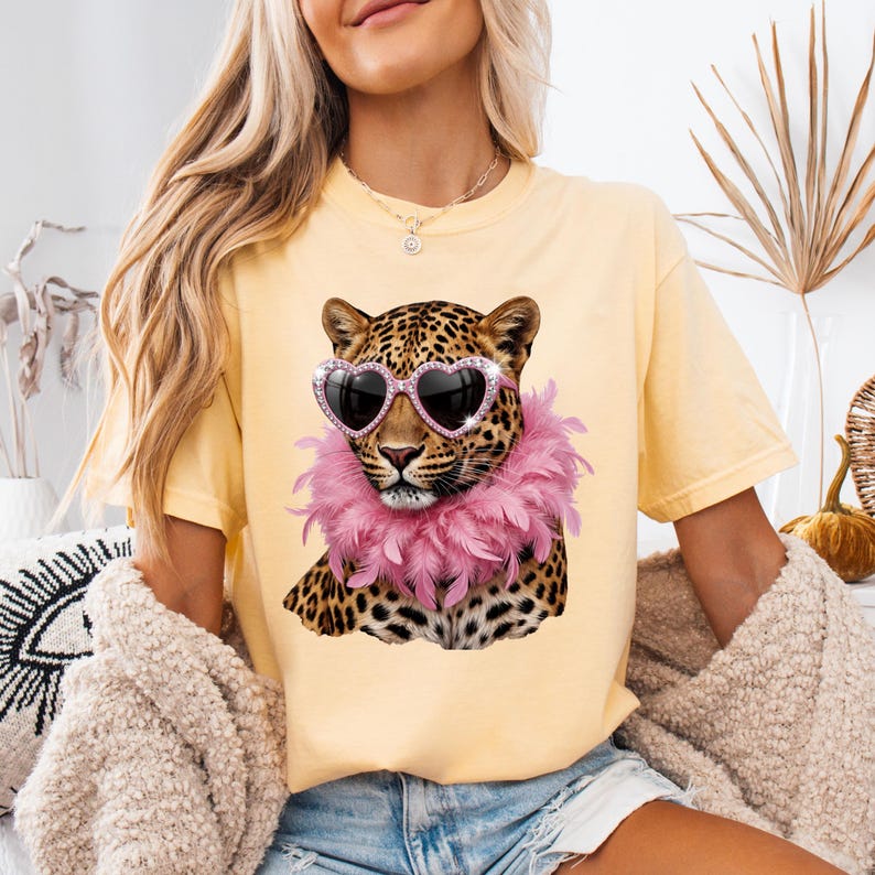May include: A pale yellow short-sleeve t-shirt featuring a graphic print of a leopard wearing pink heart-shaped sunglasses and a fluffy pink feather boa around its neck. The leopard has black spots on a tan coat and a detailed facial expression.