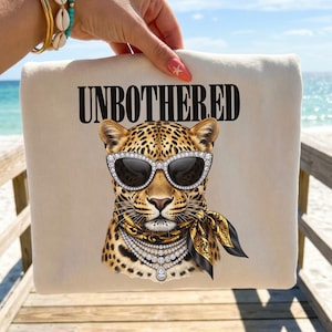 May include: A cream-colored sweatshirt featuring a graphic of a leopard wearing rhinestone-encrusted cat-eye sunglasses, a black and gold patterned neck scarf, and layered rhinestone necklaces. The text UNBOTHERED is printed in black above the leopard.