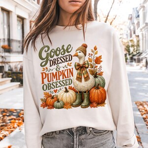 May include: A cream-colored crewneck sweatshirt featuring a graphic print of a white goose wearing a brown knit hat, a plaid scarf, and green rain boots. The goose is surrounded by orange and green pumpkins and autumn leaves with the text Goose DRESSED & PUMPKIN OBSESSED.