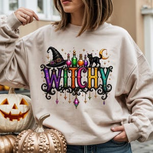 May include: A beige crewneck sweatshirt featuring a colorful graphic print with the word WITCHY in ornate, multi-colored lettering. The design includes a black witch hat, small potion bottles, a black cat, a crescent moon, and hanging crystals.