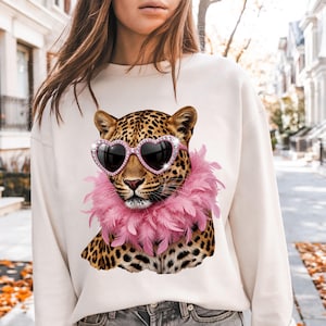 May include: A cream-colored crewneck sweatshirt featuring a graphic print of a leopard wearing pink heart-shaped sunglasses with rhinestone accents and a fluffy pink feather boa around its neck, set against a blurred outdoor street background.
