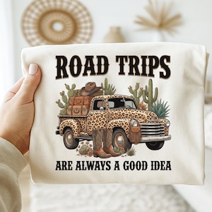 Puede incluir: Camiseta color crema con un camión vintage con estampado de leopardo, cargado de equipaje, un sombrero de vaquero y botas. El texto "ROAD TRIPS ARE ALWAYS A GOOD IDEA" está impreso arriba y abajo del camión.