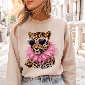 May include: A beige crewneck sweatshirt featuring a graphic print of a leopard wearing pink heart-shaped sunglasses and a pink feather boa. The leopard has a spotted coat with black and brown markings, and the design is centered on the chest.