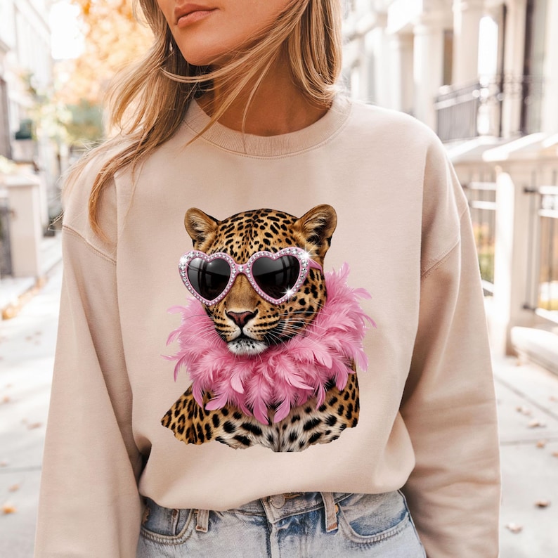 May include: A beige crewneck sweatshirt featuring a graphic print of a leopard wearing pink heart-shaped sunglasses and a pink feather boa. The leopard has a spotted coat with black and brown markings, and the design is centered on the chest.