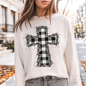 May include: A cream-colored long-sleeve sweatshirt featuring a large, central cross graphic. The cross has a black and white buffalo plaid pattern and is surrounded by a frayed, distressed-looking fabric edge detail.