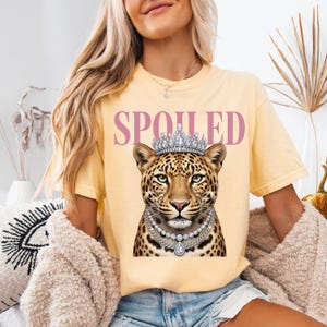 May include: A pale yellow short-sleeve t-shirt featuring a graphic of a leopard wearing a silver crown and two layered diamond necklaces. The word SPOILED is printed in large pink capital letters above the leopard illustration on the front.