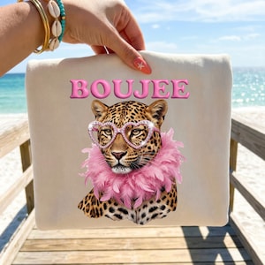 May include: A cream-colored sweatshirt featuring a graphic of a leopard wearing pink heart-shaped glasses and a pink feather boa. The word BOUJEE is printed in pink bubble letters above the leopard. The design is centered on the front of the garment.