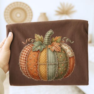 May include: A brown fabric with a patchwork pumpkin design. The pumpkin is made of various patterned fabrics in orange, yellow, green, and white, with a green and white checkered stem. Decorative leaves and swirls complete the fall-themed design.