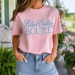 Może przedstawiać: Jasnoróżowy t-shirt z krótkim rękawem z niebieskim napisem Blue Collar BOUJEE, łączącym czcionkę skryptową i szeryfową. Koszulka jest zestawiona z jasnoniebieskimi dżinsami z widocznym srebrnym guzikiem i kieszeniami.