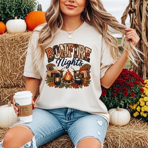 May include: Cream-colored t-shirt with the words "BONFIRE Nights" in a plaid and script design. The graphic features a campfire scene with chairs, a lantern, and fall leaves. The person is wearing blue jeans.
