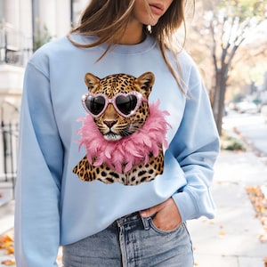 May include: A light blue crewneck sweatshirt featuring a graphic print of a leopard wearing pink heart-shaped sunglasses and a fluffy pink feather boa around its neck. The leopard has a spotted coat and is centered on the front of the garment.