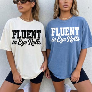 Könnte beinhalten: Zwei kurzärmelige T-Shirts mit Rundhalsausschnitt. Das linke T-Shirt ist cremefarben mit schwarzem Text FLUENT in Eye Rolls. Das rechte T-Shirt ist blau mit weißem Text FLUENT in Eye Rolls. Beide T-Shirts haben einen lockeren, weiten Schnitt.
