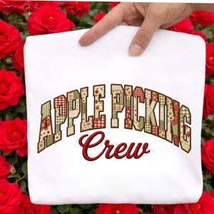 May include: A white t-shirt featuring the text APPLE PICKING in a block font with a patchwork pattern of red, green, and cream colors, and the word Crew in a dark red cursive script underneath, centered on the front of the garment.