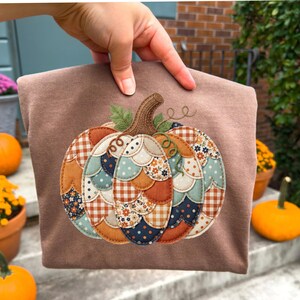 May include: A brown t-shirt featuring a large pumpkin applique made from various fabric patches. The pumpkin design uses scalloped fabric pieces in shades of orange, blue, cream, and brown with floral, polka dot, and gingham patterns.