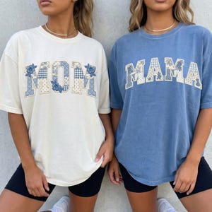 Pode incluir: Duas t-shirts de manga curta e gola redonda. A t-shirt da esquerda é creme com a palavra MOM em letras de patchwork com motivos florais e xadrez vichy azul e branco. A da direita é azul com a palavra MAMA em letras de patchwork similares.