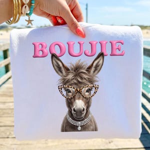 Könnte beinhalten: Ein weißes Sweatshirt mit einer Grafik eines braunen Esels, der eine Brille mit Leopardenmuster und eine mehrreihige Perlenkette trägt. Das Wort BOUJIE ist in großen, rosa, dreidimensionalen Buchstaben über dem Esel abgebildet.