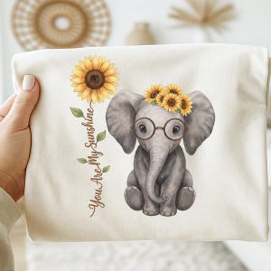 May include: A cream-colored t-shirt featuring a watercolor illustration of a gray elephant wearing round glasses and a sunflower crown. A large sunflower and the text "You Are My Sunshine" are also printed on the shirt.