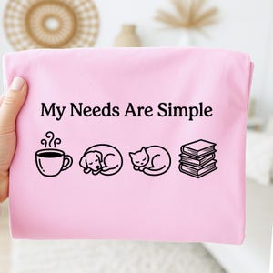 May include: A light pink t-shirt with the text "My Needs Are Simple" in black. Below the text are black line drawings of a coffee cup, a sleeping dog, a sleeping cat, and a stack of books.