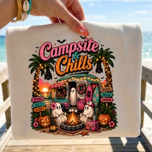 May include: Cream-coloured sweatshirt with a colourful graphic featuring a campsite scene with ghosts, a campfire, and palm trees. The text "Campsite & Chills" is displayed above the scene. The design includes pumpkins, surfboards, and a sunset.