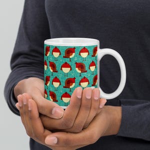May include: White ceramic mug featuring a repeating pattern of stylized red and brown birds on a teal background. The birds have white bellies and are set against a backdrop of musical notes. The mug has a curved handle.