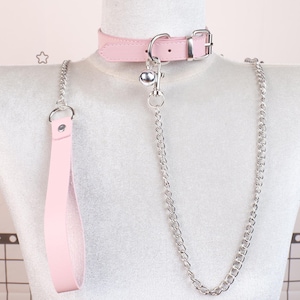 Könnte beinhalten: Ein rosa Kunstleder-Halsband und Leinen-Set. Das Halsband hat eine silberne Schnalle und eine kleine Glocke. Eine silberne Kette verbindet das Halsband mit einer passenden rosa Leine. Das Set wird auf einer Schaufensterpuppe präsentiert.
