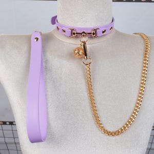 May include: A lilac collar and lead set with gold-coloured hardware. The collar has a buckle closure and decorative rivets. A gold chain and charm are attached to the collar. The lead is a solid lilac colour.