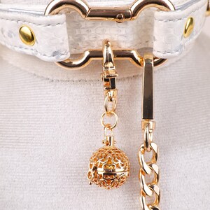 May include: White faux leather collar with gold-tone hardware, including a chain and a decorative filigree ball charm. The collar features a textured pattern and gold rivets. The chain and charm are suspended from a gold clasp.