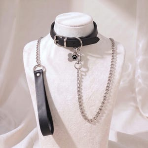 May include: A black leather collar with a silver-toned buckle and a silver metal D-ring. Attached to the D-ring is a bone-shaped silver tag featuring a black paw print design and a long silver chain leash with a black leather handle loop.